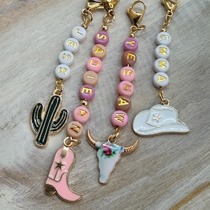 May include: Four beaded keychains with gold-coloured hardware. Each keychain has a name spelled out with letter beads. Charms include a cactus, a pink cowboy boot, a floral skull, and a white cowboy hat.
