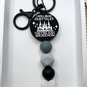 Peut inclure: Porte-cl&eacute;s noir et blanc avec un design de ch&acirc;teau et de livre. Le porte-cl&eacute;s dit "Je lis toujours des contes de f&eacute;es, ils sont juste plus &eacute;pic&eacute;s maintenant". Le porte-cl&eacute;s a un charme de perle en silicone noir et gris.