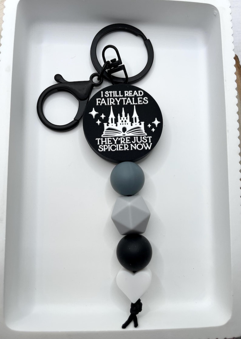 Peut inclure: Porte-cl&eacute;s noir et blanc avec un design de ch&acirc;teau et de livre. Le porte-cl&eacute;s dit "Je lis toujours des contes de f&eacute;es, ils sont juste plus &eacute;pic&eacute;s maintenant". Le porte-cl&eacute;s a un charme de perle en silicone noir et gris.