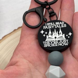 Peut inclure: Porte-cl&eacute;s noir avec un charme rond qui dit "Je lis toujours des contes de f&eacute;es, ils sont juste plus &eacute;pic&eacute;s maintenant". Le charme pr&eacute;sente un ch&acirc;teau blanc et un livre avec des &eacute;toiles. Le porte-cl&eacute;s a &eacute;galement trois perles en silicone noires et grises.