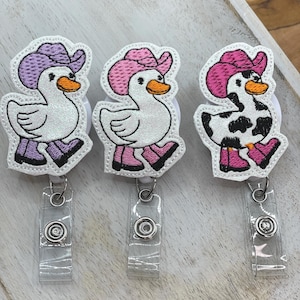 May include: Three duck-shaped badge reels with cowboy hats. The ducks have white bodies, orange beaks, and are wearing purple, pink, and pink-and-black hats. Each reel has a clear plastic strap and a metal clip.