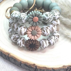 May include: A collection of beaded bracelets with floral accents. The bracelets feature a mix of sage green, white with brown leaf patterns, and flower-shaped beads in shades of pink and brown. Gold-colored key rings are also visible.