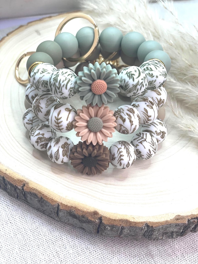 May include: A collection of beaded bracelets with floral accents. The bracelets feature a mix of sage green, white with brown leaf patterns, and flower-shaped beads in shades of pink and brown. Gold-colored key rings are also visible.