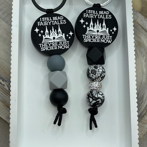 Peut inclure: Deux porte-cl&eacute;s noirs et blancs avec le texte "I still read fairytales they're just spicier now". Un porte-cl&eacute;s a un motif de ch&acirc;teau et l'autre a un motif d'&eacute;toile. Les deux porte-cl&eacute;s ont des perles noires et grises.