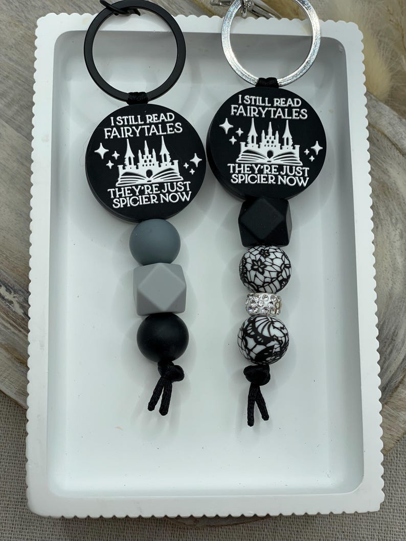 Peut inclure: Deux porte-cl&eacute;s noirs et blancs avec le texte "I still read fairytales they're just spicier now". Un porte-cl&eacute;s a un motif de ch&acirc;teau et l'autre a un motif d'&eacute;toile. Les deux porte-cl&eacute;s ont des perles noires et grises.