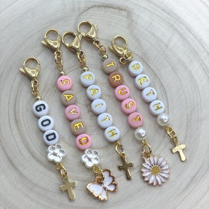 Puede incluir: Cinco llaveros de cuentas con cierres y dijes dorados. Las cuentas son blancas, rosas y beige, deletreando palabras como "GOD" y "TRUST". Los dijes incluyen cruces, una mariposa y una margarita. El fondo es de vetas de madera clara.