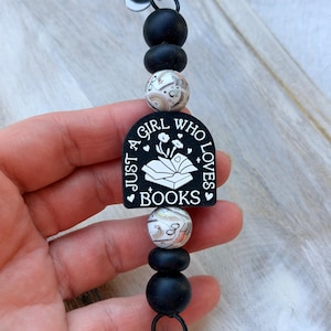May include: Black beaded keychain with a black and white graphic that reads "Just a girl who loves books." The keychain has a black clasp and decorative beads in black and white.