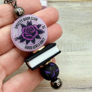 May include: A retractable badge reel featuring a lavender charm with the text "Smutty Book Club" and a purple rose design. It includes a small black book charm and black and purple bead accents.