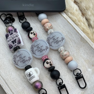 Puede incluir: Tres cordones con cuentas con mosquetones negros y varios dijes. Los dijes incluyen un libro en miniatura, una calavera y una taza de café. Los cordones tienen las frases "Good Girls Read Bad Books", "When I die, delete my Kindle history" y "Go Away I'm Reading".