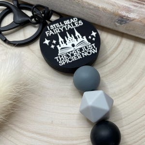 Peut inclure: Porte-cl&eacute;s en silicone noir et blanc avec un charme qui dit "Je lis toujours des contes de f&eacute;es, ils sont juste plus &eacute;pic&eacute;s maintenant". Le charme repr&eacute;sente un ch&acirc;teau et un livre. Le porte-cl&eacute;s comprend &eacute;galement un ensemble de perles en silicone noir, gris et blanc.