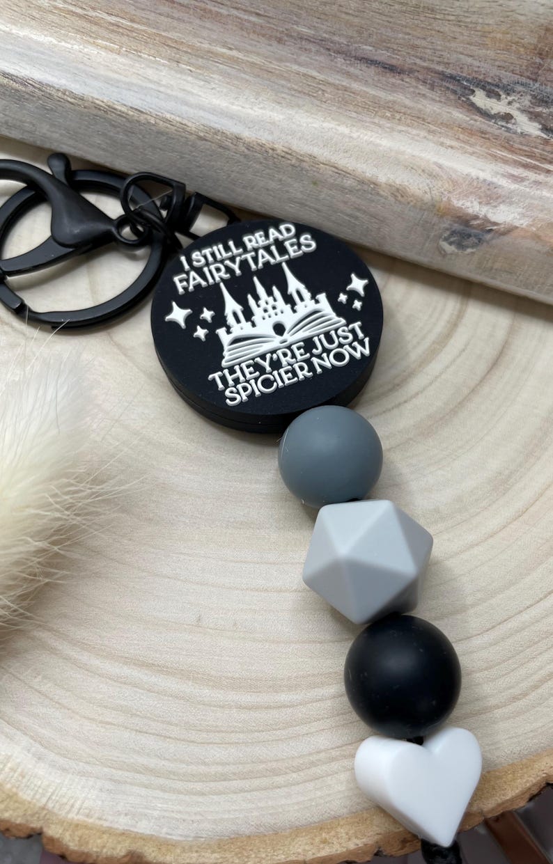 Peut inclure: Porte-cl&eacute;s en silicone noir et blanc avec un charme qui dit "Je lis toujours des contes de f&eacute;es, ils sont juste plus &eacute;pic&eacute;s maintenant". Le charme repr&eacute;sente un ch&acirc;teau et un livre. Le porte-cl&eacute;s comprend &eacute;galement un ensemble de perles en silicone noir, gris et blanc.