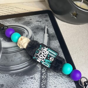 Pu&ograve; includere: Un cordino a tema "Spooky Book Club" in bianco e nero con una perlina a forma di teschio, perline turchesi e viola e un accento nero e soffice. Il cordino &egrave; sopra un e-reader.