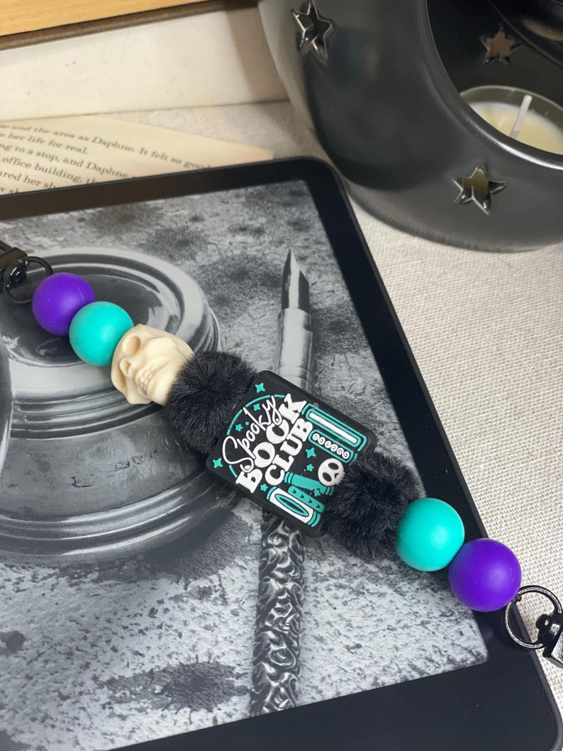 Pu&ograve; includere: Un cordino a tema "Spooky Book Club" in bianco e nero con una perlina a forma di teschio, perline turchesi e viola e un accento nero e soffice. Il cordino &egrave; sopra un e-reader.