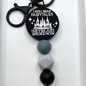 Peut inclure: Porte-cl&eacute;s noir et blanc avec un charme qui dit "Je lis toujours des contes de f&eacute;es, ils sont juste plus &eacute;pic&eacute;s maintenant". Le charme repr&eacute;sente un ch&acirc;teau et un livre. Le porte-cl&eacute;s a &eacute;galement une cha&icirc;ne de perles en silicone noir et blanc avec un charme en forme de c&oelig;ur blanc.