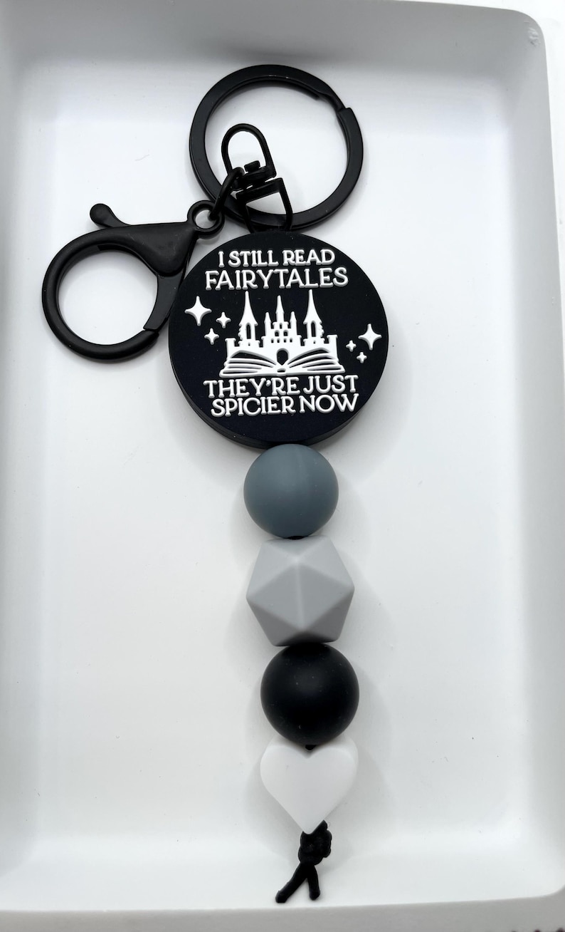 Peut inclure: Porte-cl&eacute;s noir et blanc avec un charme qui dit "Je lis toujours des contes de f&eacute;es, ils sont juste plus &eacute;pic&eacute;s maintenant". Le charme repr&eacute;sente un ch&acirc;teau et un livre. Le porte-cl&eacute;s a &eacute;galement une cha&icirc;ne de perles en silicone noir et blanc avec un charme en forme de c&oelig;ur blanc.