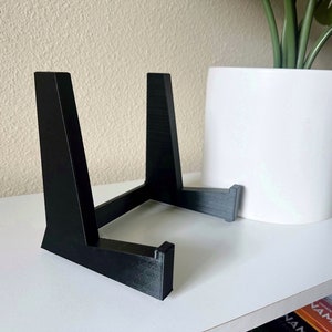 May include: A black, angled plant stand with a three-legged design. The stand is made of a solid material and is designed to hold a potted plant. The stand is approximately 8 inches tall and 10 inches wide.