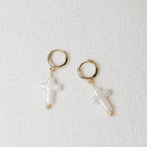 May include: A pair of gold hoop earrings with a white pearl cross charm hanging from each hoop.