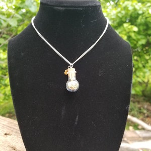 May include: A silver chain necklace with a small glass bottle pendant filled with gold glitter. The bottle has a cork stopper with a small gold charm attached.