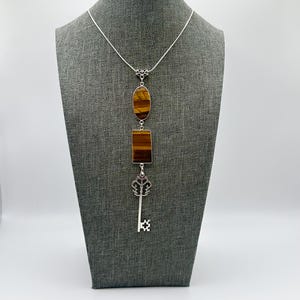 May include: A silver necklace with a pendant featuring an oval and rectangular tiger's eye stone, and a decorative key charm. The necklace is displayed on a gray mannequin.