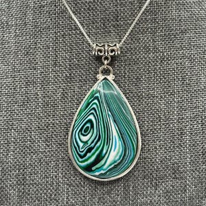 May include: A teardrop-shaped pendant necklace featuring a swirling pattern of dark green, light green, teal, and white colors. The pendant is encased in a silver-toned metal frame with an ornate bail and hangs from a thin silver-toned chain.