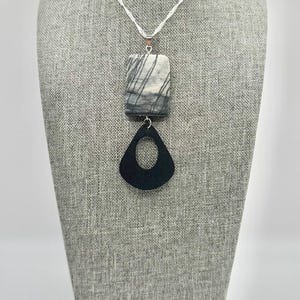 May include: A necklace featuring a silver-toned chain with a pendant consisting of a rectangular gray and white stone piece with dark linear veining, attached to a black teardrop-shaped component with a central cutout, creating a layered design.