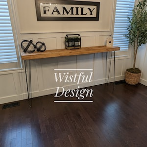 Peut inclure: Une longue et étroite console en bois avec des pieds en épingle noirs. La table est ornée d'objets décoratifs et du mot "FAMILY" dans un cadre au-dessus. L'image présente également le texte "Wistful Design".