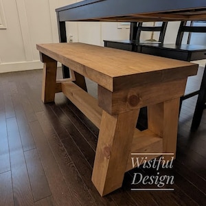 May include: A rustic wooden bench with a natural wood finish. The bench has a simple design with a wide seat and two sturdy legs. The bench is made of solid wood and has a natural wood grain. The bench is perfect for adding a touch of rustic charm to any room.