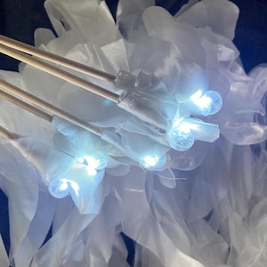 May include: Close-up of illuminated white floral decorations with wooden dowels. Each flower has a bright LED light at its center, creating a soft glow. The background is a dark blue, enhancing the brightness of the lights and the white fabric.