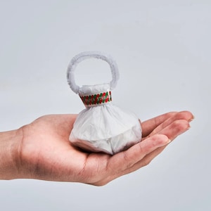 May include: A small, white paper streamer popper with a white handle and a decorative band that reads "PAPER STREAM". The popper is held in an open hand against a white background. The band has red and green accents.