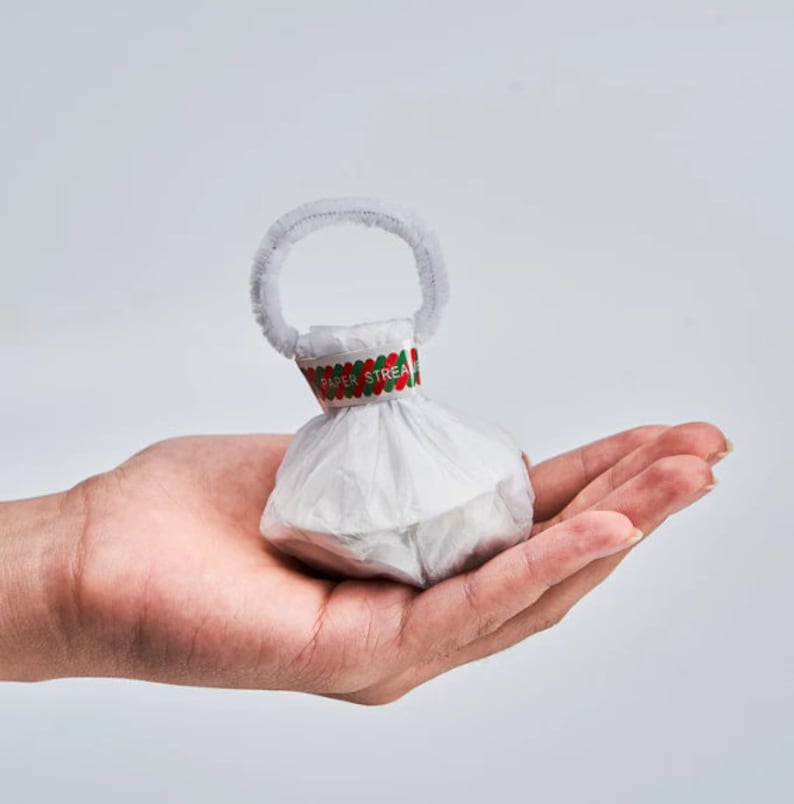 May include: A small, white paper streamer popper with a white handle and a decorative band that reads "PAPER STREAM". The popper is held in an open hand against a white background. The band has red and green accents.
