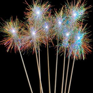 May include: A collection of seven light-up wands featuring iridescent, holographic tinsel pom-poms attached to thin, light-colored wooden sticks. Each pom-pom emits a bright white LED glow from the center, creating a starburst effect with colorful light reflections across the metallic strands.