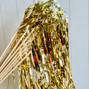 May include: Gold streamer wands with white plastic balls at the top. The wands are made of wooden dowels and have gold foil streamers attached.