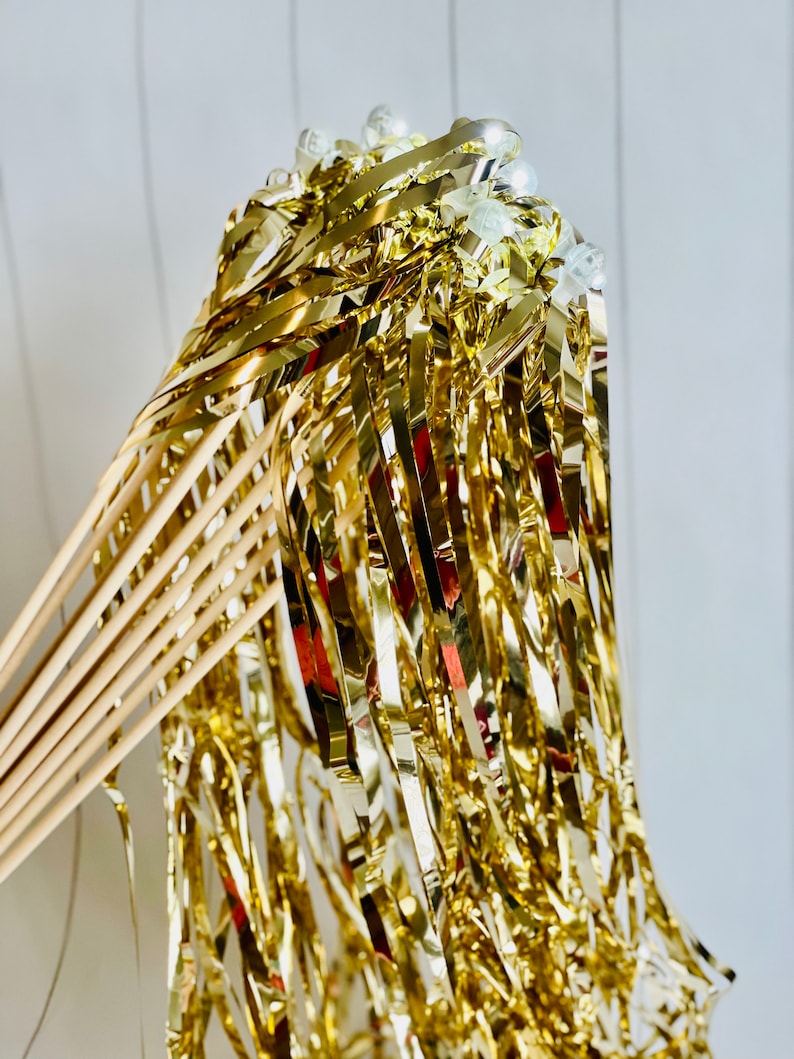 May include: Gold streamer wands with white plastic balls at the top. The wands are made of wooden dowels and have gold foil streamers attached.
