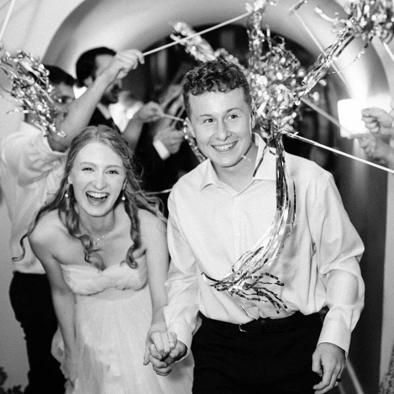 May include: A bride and groom smile as they walk through a doorway decorated with silver streamers. The bride is wearing a white strapless dress and the groom is wearing a white button-down shirt and black pants.