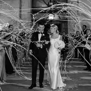 May include: Black and white photograph of a wedding celebration. A couple, the bride in a white gown and the groom in a tuxedo, walk through a shower of streamers. Guests line the sides, holding the streamers aloft.