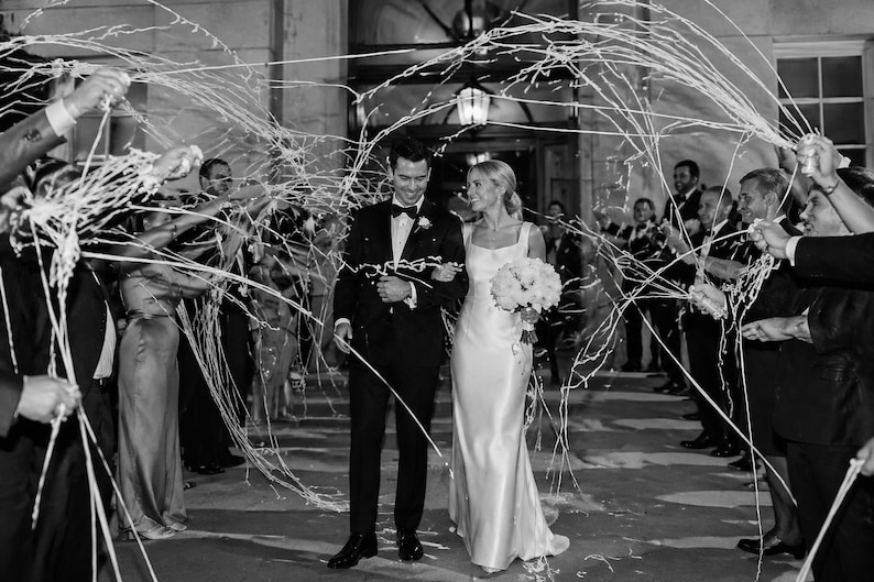 May include: Black and white photograph of a wedding celebration. A couple, the bride in a white gown and the groom in a tuxedo, walk through a shower of streamers. Guests line the sides, holding the streamers aloft.