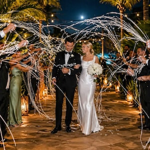 May include: A newly married couple walks down a path lined with guests throwing confetti. The bride wears a white satin gown and carries a bouquet of white flowers. The groom is in a black tuxedo. The scene is lit by string lights and a full moon.