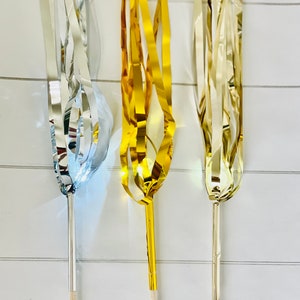 May include: Three streamer wands with wooden sticks. The wands are made of silver, gold, and champagne colored metallic streamers.