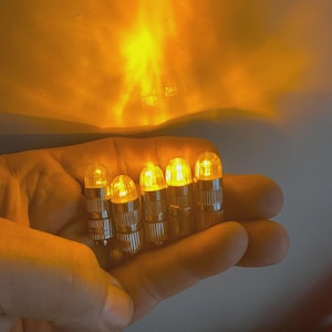 May include: Five small, battery-operated LED lights with a warm yellow glow. Each light has a clear, rounded top and a silver base. The lights are held in a hand, with a soft, diffused light in the background.