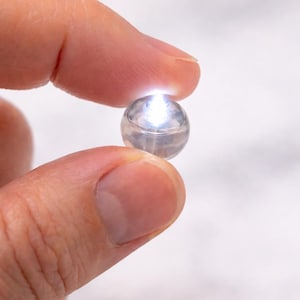 May include: A small, clear, spherical object with a bright, white light emanating from within. The object is held between two fingers against a soft, blurred background. The light source appears to be contained within the sphere.
