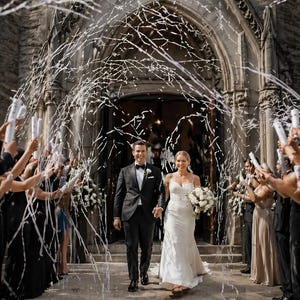 May include: A newly married couple exits a stone church, showered with white confetti. The bride wears a white wedding dress and carries a bouquet. The groom is in a black tuxedo. Guests hold confetti cannons, celebrating the wedding.