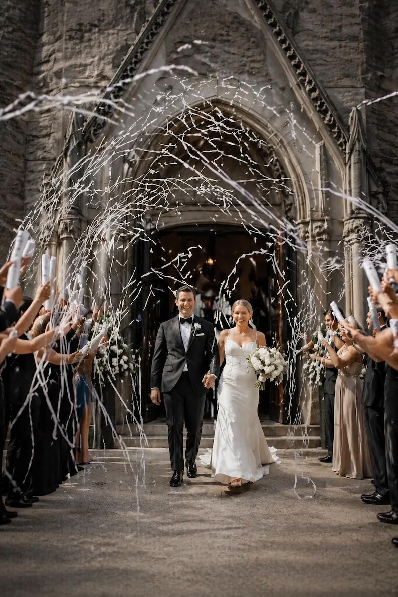 May include: A newly married couple exits a stone church, showered with white confetti. The bride wears a white wedding dress and carries a bouquet. The groom is in a black tuxedo. Guests hold confetti cannons, celebrating the wedding.