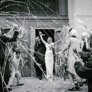 May include: Black and white photograph of a wedding celebration. A bride and groom exit a building, surrounded by guests throwing streamers. The bride holds a bouquet, and the groom wears a dark suit. The image captures a moment of joy and festivity.