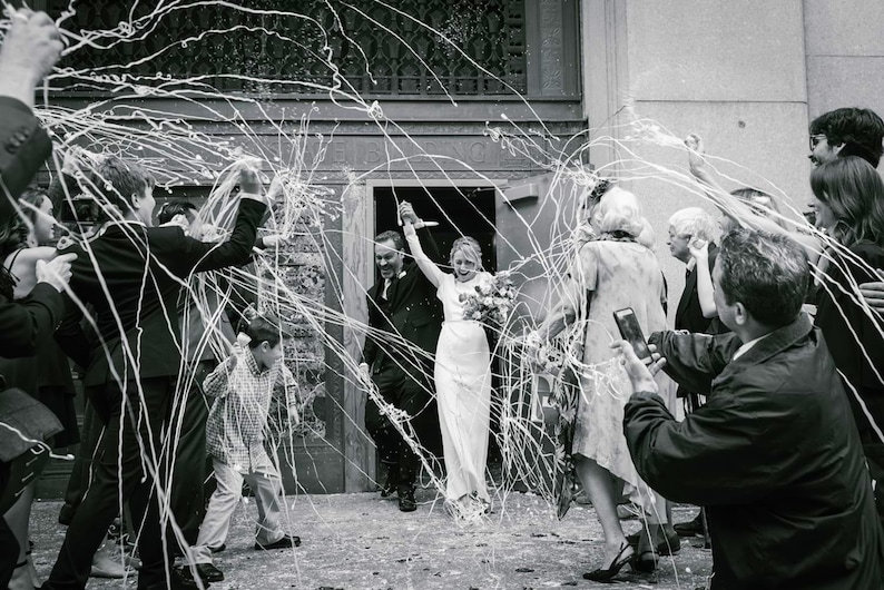 May include: Black and white photograph of a wedding celebration. A bride and groom exit a building, surrounded by guests throwing streamers. The bride holds a bouquet, and the groom wears a dark suit. The image captures a moment of joy and festivity.