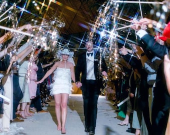 May include: A newly married couple walks through a tunnel of sparklers held by wedding guests. The bride wears a white, feathered mini-dress and a cowboy hat. The groom is in a black tuxedo. The scene is illuminated by sparkling lights.