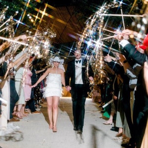 May include: A bride and groom walk down a pathway, surrounded by guests holding sparklers, as they exit their wedding ceremony.