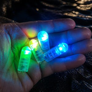 May include: Four small, clear LED lights in green, yellow, and blue are held in a hand. The lights are cylindrical with rounded tops and are illuminated, creating a colorful glow. The background is dark.