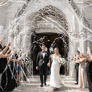 May include: A newly married couple exits a church, showered with white confetti. The bride wears a white gown and carries a bouquet. The groom is in a black tuxedo. Guests hold confetti cannons, creating a celebratory atmosphere.