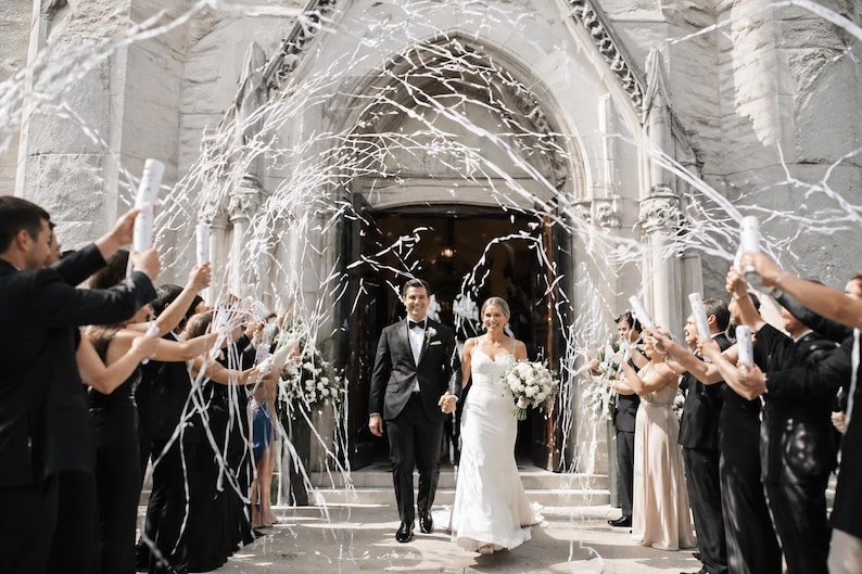 May include: A newly married couple exits a church, showered with white confetti. The bride wears a white gown and carries a bouquet. The groom is in a black tuxedo. Guests hold confetti cannons, creating a celebratory atmosphere.