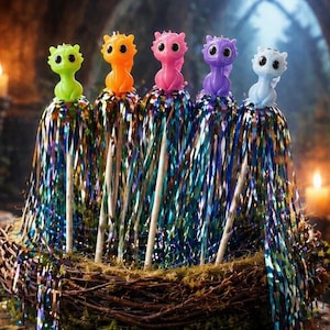 Puede incluir: Una exhibición caprichosa con cinco figuras de dragones coloridos, verde, naranja, rosa, morado y blanco, sobre varitas decorativas. Las varitas están adornadas con serpentinas brillantes multicolores, colocadas en un nido rústico.